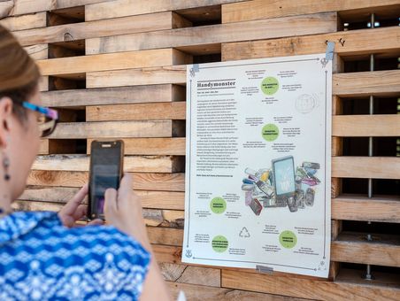 An einer Holzwand hängt ein Plakat über "Handymonster". Eine Frau fotografiert es.