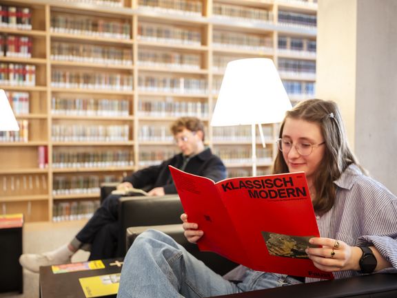 Zwei Personen lesen entspannt in Sesseln in einer gemütlichen, hellen Sitzecke in der Bibliothek.
