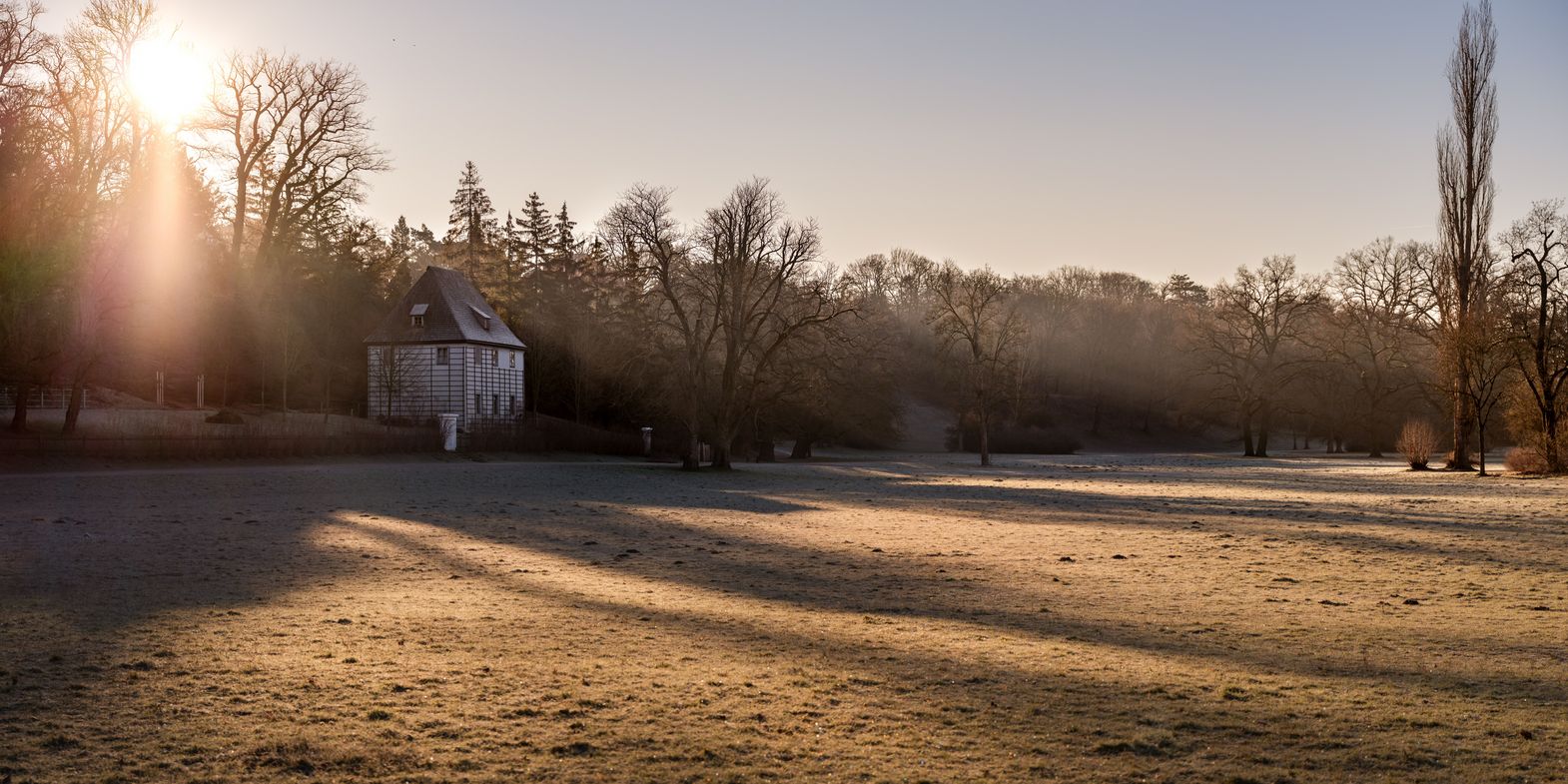 Ein sonniges Feld im Park an der Ilm mit Goethes Gartenhaus im Hintergrund. Die Sonne scheint von links und wirft lange Schatten auf das leicht gefrorene Gras. Im Hintergrund sind kahle Bäume und ein bewaldetes Gebiet zu sehen.