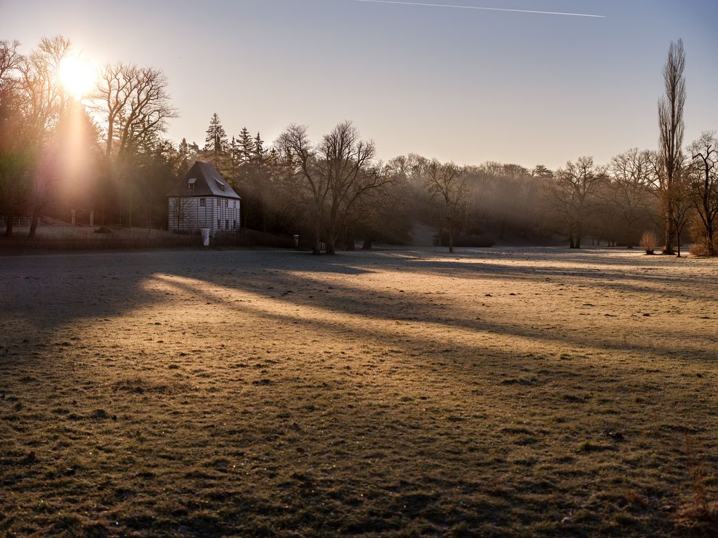 Ein sonniges Feld im Park an der Ilm mit Goethes Gartenhaus im Hintergrund. Die Sonne scheint von links und wirft lange Schatten auf das leicht gefrorene Gras. Im Hintergrund sind kahle Bäume und ein bewaldetes Gebiet zu sehen.