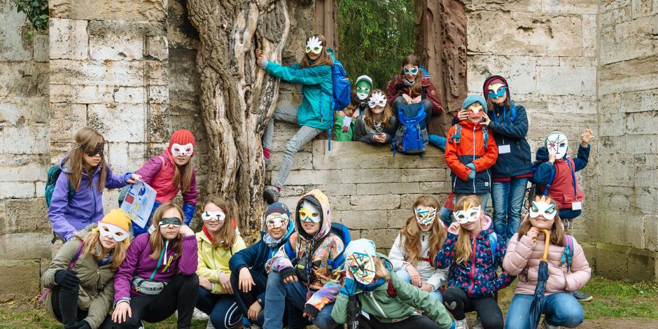 Junge Schulklasse posiert mit selbstgebastelten Masken für ein Gruppenfoto. Im Hintergrund eine Ruine.