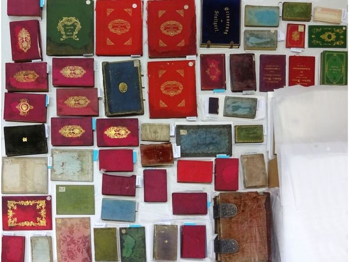 Zu sehen sind fünfzig unterschiedlich große Bücher mit Seiden- und Seidensamteinbänden, die durch Hitze und Löschwasser beschädigt wurden. Die Fotografie zeigt die Einbände vor der Restaurierung.