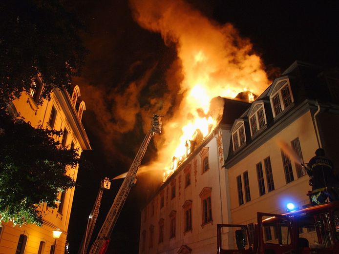 Auf der Fotografie sieht man, wie der Dachstuhl des Historischen Gebäudes der Herzogin Anna Amalia Bibliothek in der Nacht vom 2. Septembers 2004 brennt. Auf zwei Feuerleitern und einem Einsatzwagen sind Feuerwehrmänner dabei, den Brand zu löschen.