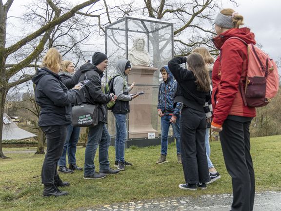 Eine Gruppe Jugendlicher testet die Rallye auf Ipads. Sie stehen im Park vor einer Büste.