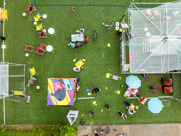 Eine Drohnenaufnahme blickt von oben auf einen grünen künstlichen Rasen mit Fußballtor, Leinwand und Sitzgelegenheiten. Es erinnert an ein großes Wimmelbild. 