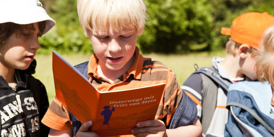 A boy standing in the park with his friends, reading a tour booklet aloud