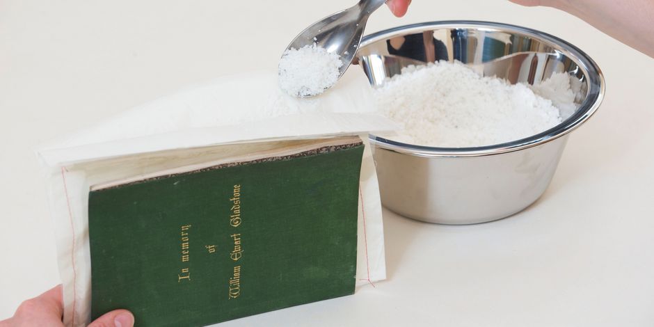 Zu sehen ist ein Buch, das einen durch Löschwasser deformierten Gewebeeinband hat. Mit Hilfe einer Arbocel-Kompresse, die das Material indirekt befeuchtet, wird die Deformierung reduziert.