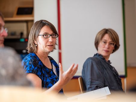Eine Frau mit blauem Oberteil erklärt etwas. Sie sitzt zwischen anderen Personen.