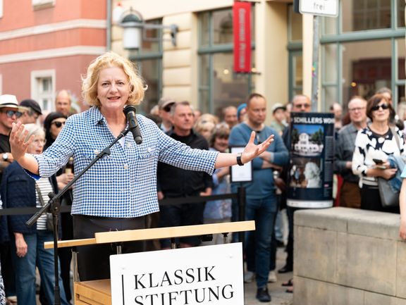 Eine Person mit karierter Bluse spricht in ein Mikron und hält die Hände nach oben.