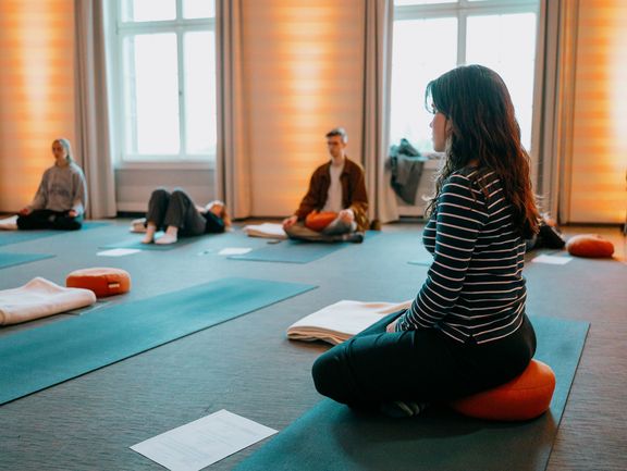 Drei Menschen sitzen auf Matten und Sitzkissen im Schneidersitz.