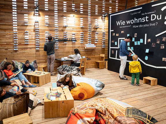 Im Innenraum sitzen einige Personen auf gemütlichen Sitzkissen und sind konzentriert am Basteln oder am Lesen. Ein Vater und sein Sohn stehen vor einer etwa drei Meter hohen Beteiligungswand zur Frage "Wie wohnst Du?". Darauf wurden verschiedene handgeschriebene Zettel von Teilnehmenden angebracht.