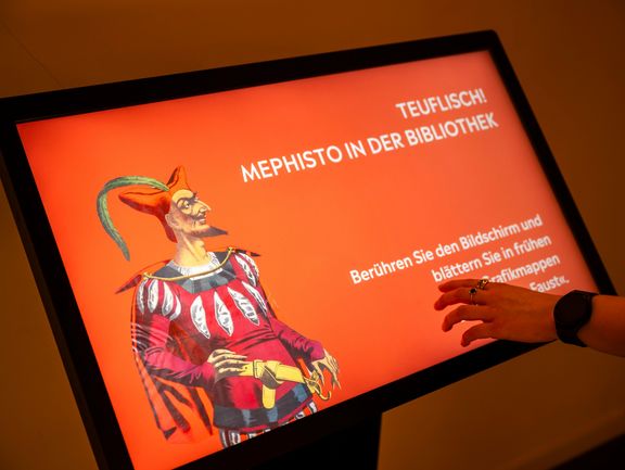 Eine Hand berührt einen roten Bildschirm auf dem links im Bild ein Teufel zu erkennen ist.