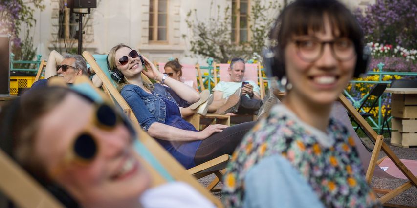 Entspannte junge Menschen liegen mit Kopfhörern auf den Ohren auf Liegestühlen vor dem Schloss.