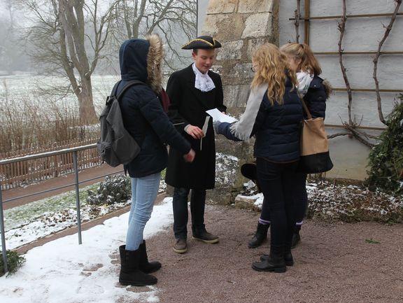 Young people at the Goethe Gartenhaus in the winter. A boy is wearing a historic costume and holding a quill. The others are reading a text.