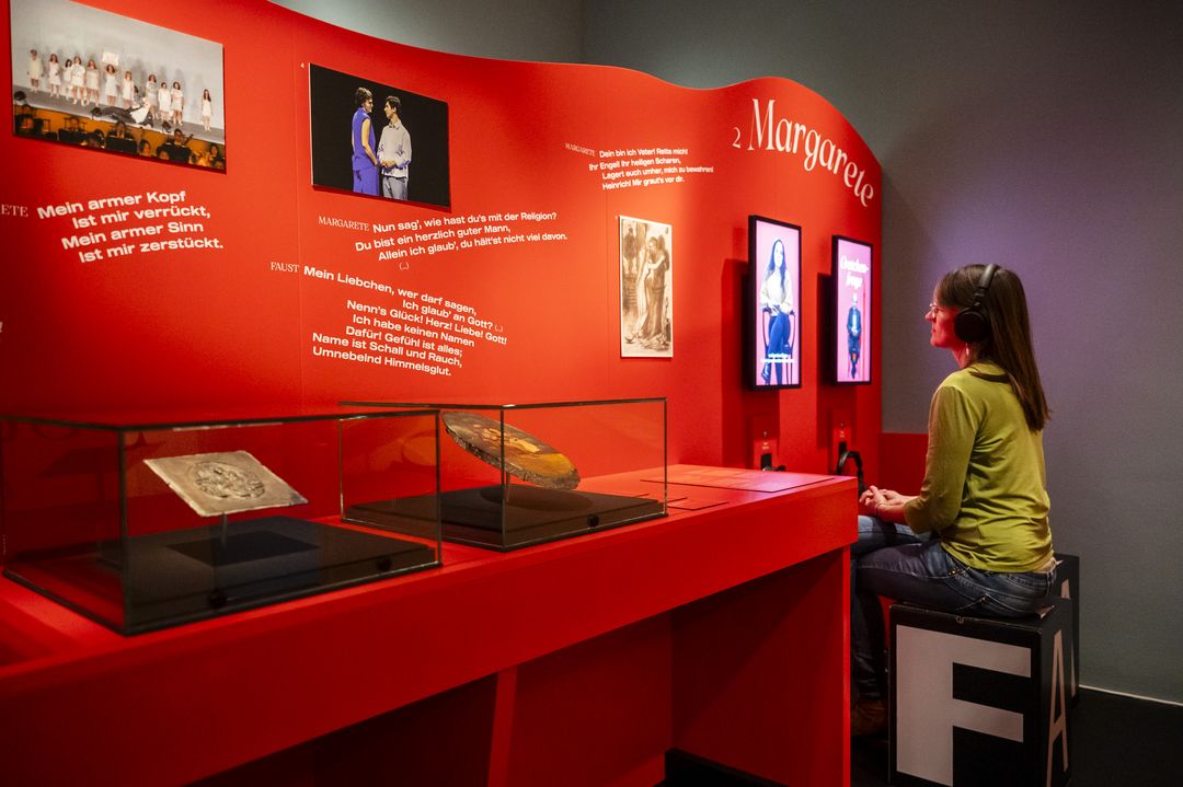 Eine große rote Wand mit Texten, Grafiken und Objekten im Glaskasten