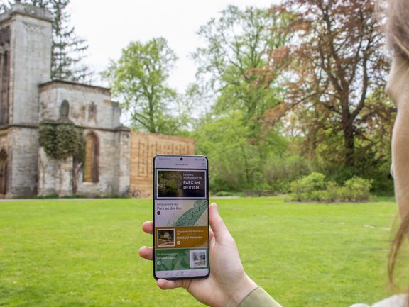 A woman, facing the Tempelherrenhaus, holds a smartphone with the Weimar+ app opened