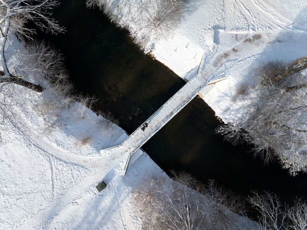 Blick von oben auf eine weiße Schneelandschaft mit einer Brücke über einen langen dunklen Fluss.