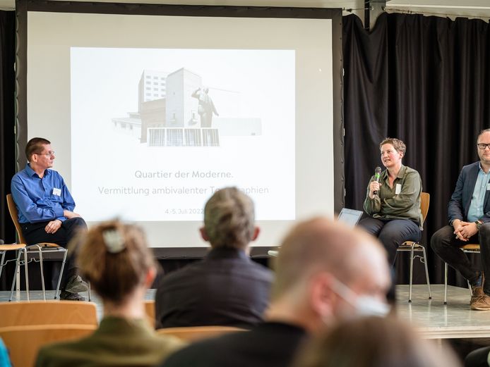 Auf einer Bühne sitzen drei Personen, eine hält ein Mikrofon. Im Hintergrund steht eine Leinwand mit dem Logo und dem Titel der Tagung