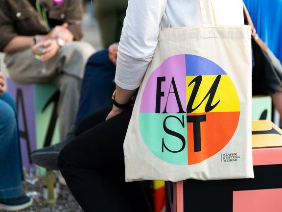 Eine Person hält eine Stofftasche mit dem Aufdruck "FAUST" in verschiedenen Farben.