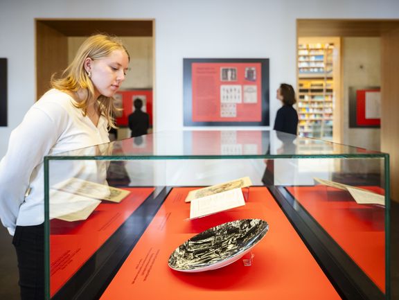 Eine junge Frau betrachtet Ausstellungsstücke in einer Vitrine. Darunter ein schwarzer Teller mit Mephisto-Zeichnung.