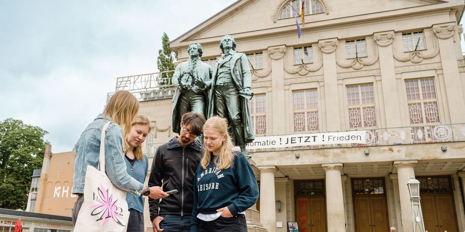 Vor dem Goethe- und Schiller-Denkmal steht eine Gruppe von vier Personen, die gebannt auf ein Smartphone schauen und Inhalte der Kulturapp abspielen.