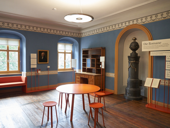 Blaugestrichener Raum, der Tisch und Stühle in der Raummitte, an den Wänden einen Ofen, einen Schreibtisch und viele auf Stäben fixierten Texttafeln beinhaltet