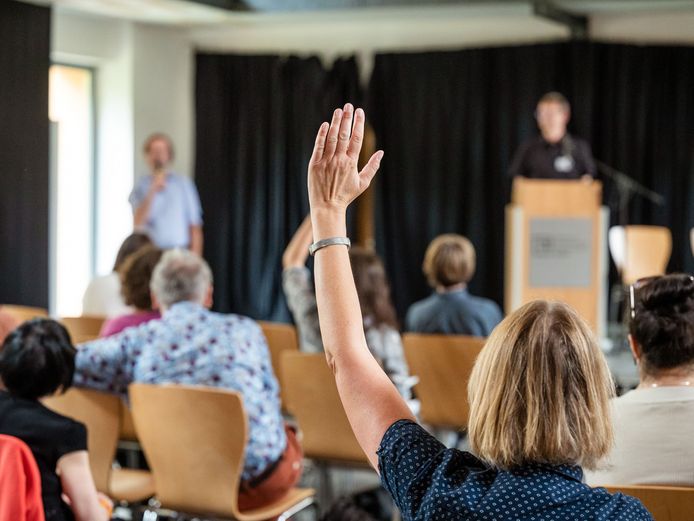 Eine Frau aus dem Publikum hebt die Hand. Sie blickt auf ein Podium und einen Moderator.