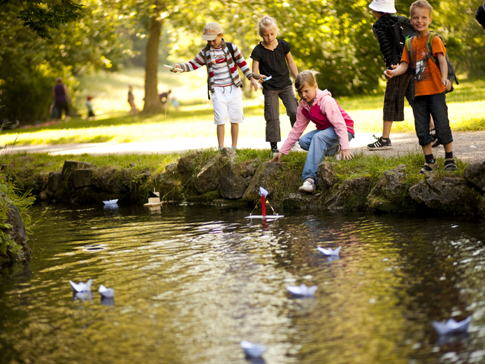 Kinder stehen an einem Fluss und sehen zu, wie Papierboote auf dem Wasser schwimmen.