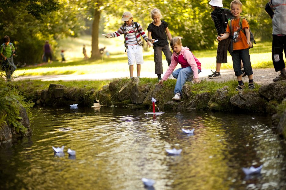 Kinder stehen an einem Fluss und sehen zu, wie Papierboote auf dem Wasser schwimmen.