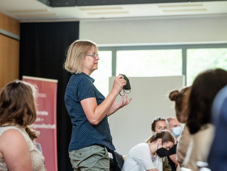 Eine Frau aus dem Publikum spricht. Sie steht, während andere Teilnehmende neben ihr sitzen.