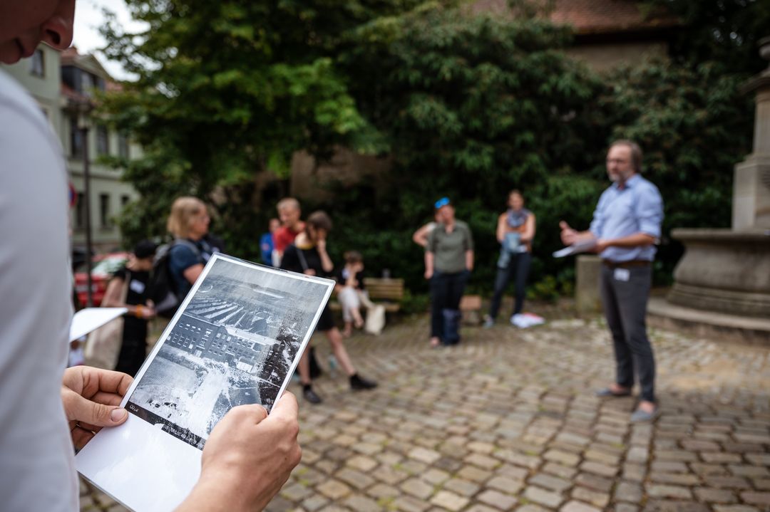 Ein Stadtführer erklärt etwas. Im vorderen Bildausschnitt betrachtet einer der Zuhörenden ein historisches Foto.