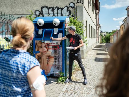 Ein Mann steht an einem Altkleidercontainer, an dem zwei große Augen und Zähne aufgeklebt sind.
