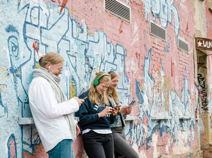 A group of four people, standing in front of a graffiti-covered wall, listen and watch attentively to the content of the cultural app on a smartphone