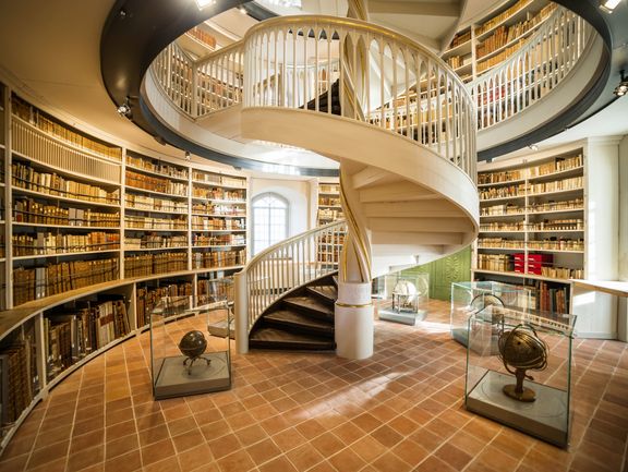 In einem Raum mit kreisförmigen Grundriss sind um eine spiralförmige Treppe mehrere Globen aufgestellt. Die Treppe führt in die oberen Etagen des Turmgebäudes, in dem Bücher aufbewahrt werden.