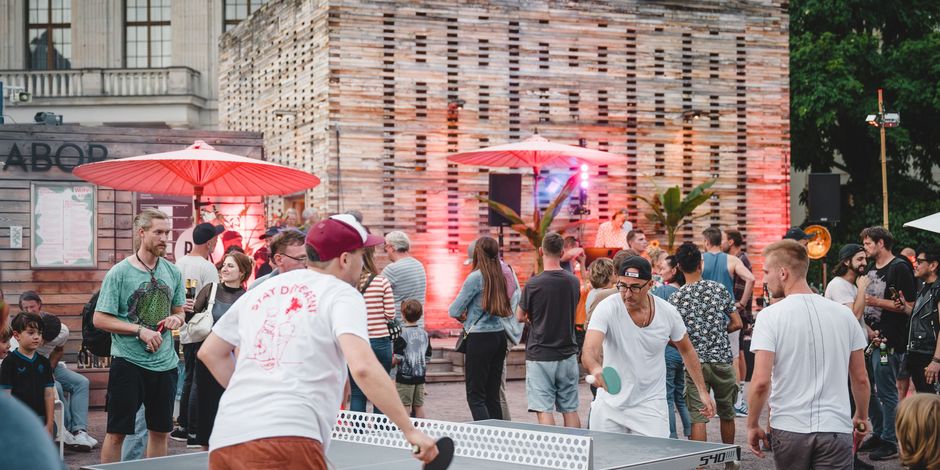 Zwei Menschen spielen Tischtennis, im Hintergrund sind Gäste einer Veranstaltung vor dem Co-Labor zu sehen