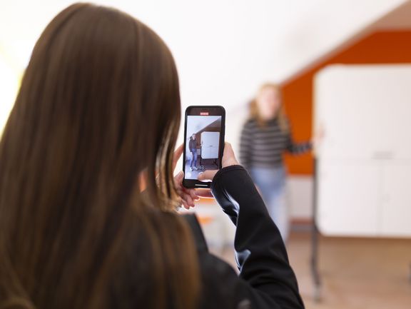 Eine Person macht mit ihrem Smartphone ein Foto. Im Hintergrund präsentiert eine Schülerin einen Schrank.