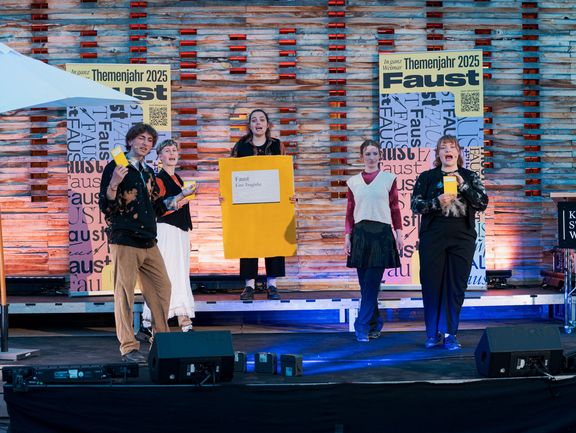 Fünf Personen stehen auf einer Bühne. Die dritte Person von links trägt ein großes, gelbes Reclam-Heft mit dem Titel "Faust. Eine Tragödie" als Kostüm.