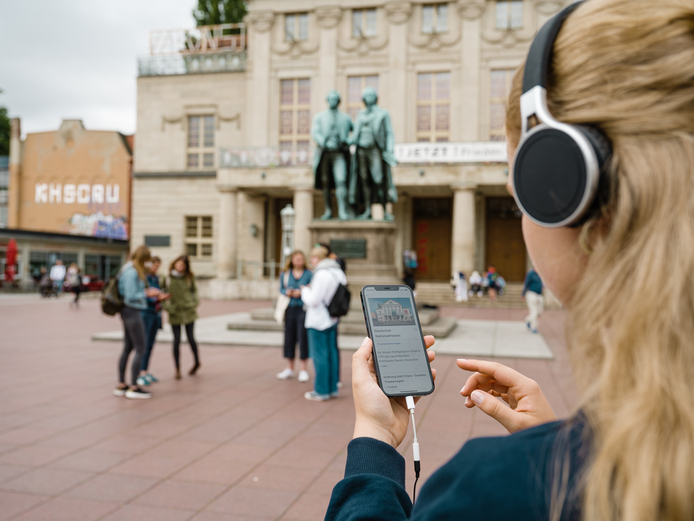Frau lauscht über die Kopfhörer einer Hörgeschichte zum Deutschen Nationaltheater, ihr Blick geht Richtung Goethe- und Schiller-Denkmal