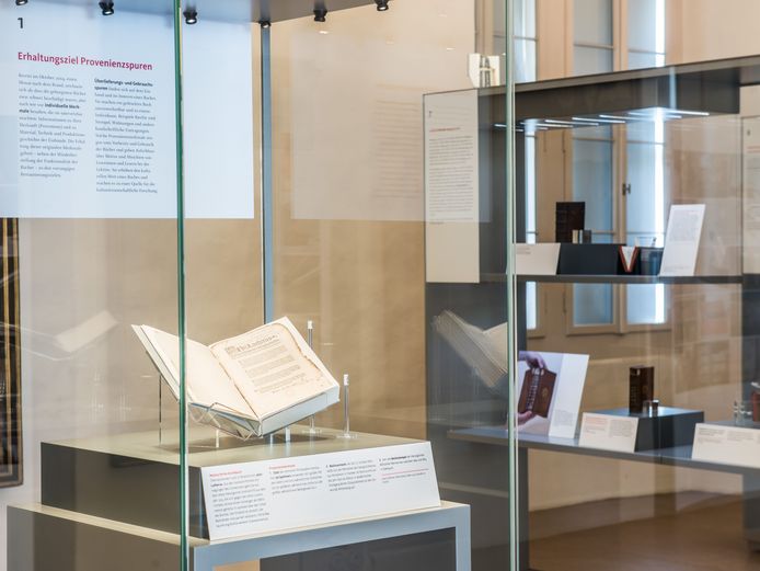 Auf der Fotografie sieht man zwei Vitrinen aus der Ausstellung "Restaurieren nach dem Brand - Die Rettung der Bücher der Herzogin Anna Amalia Bibliothek". In der vorderen Vitrine steht ein restauriertes Aschebuch aufgeschlagen.