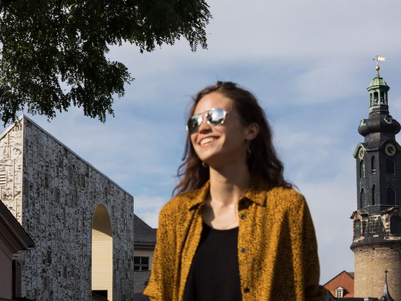 Im Vordergrund steht eine Frau mit Sonnenbrille, die Freude ausstrahlt und im Hintergrund befinden sich ein Schlossturm und einem Neubau aus mehrschichtigen Massivholzplatten