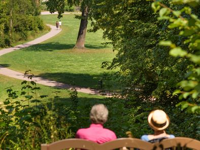 Zwei Parkbesucher genießen den Ausblick von einer Parkbank auf Goethes Gartenhaus.