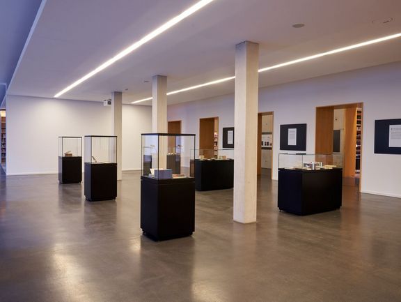 Sechs schwarze Vitrinen mit Glashauben, die rechts uns links von drei weißen Säulen aufgestellt sind. An der rechten Wand sind große Durchgänge zum Bücherkubus. Dazwischen hängen schwarze Tafeln mit Erklärungen zu den in den Vitrinen ausgestellten Objekten.