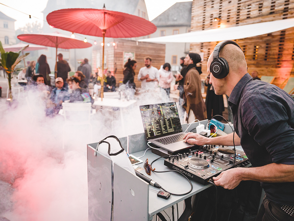 Ein DJ legt vor dem festlich bedeuteten Co-Labor auf. Davor unterhalten sich in Nebel gehüllt mehrere Personen