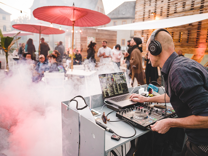 Ein DJ legt vor dem festlich bedeuteten Co-Labor auf. Davor unterhalten sich in Nebel gehüllt mehrere Personen