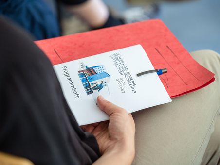 Auf dem Schoß einer Teilnehmerin liegt eine rote Mappe, ein Kugelschreiber und das Programmheft der Tagung.