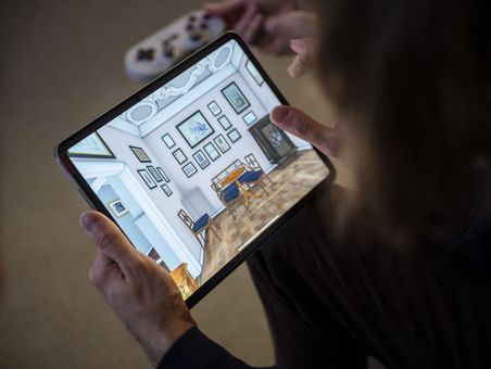 Goethe-Live-3D Eine Person hält ein Tablet in den Händen, auf dessen Bildschirm ein virtuelles Zimmer zu sehen ist. Das Zimmer auf dem Bildschirm ist modern eingerichtet, mit weißen Wänden, die mit mehreren gerahmten Bildern dekoriert sind. In der Mitte des Raumes steht ein Tisch mit zwei blauen Stühlen. Der Boden ist mit einem hellen Holzparkett ausgelegt. Im Hintergrund ist eine Treppe zu sehen, die nach oben führt. Die Person, die das Tablet hält, ist nur teilweise sichtbar, hauptsächlich die Hände und ein Teil des Oberkörpers.