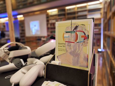 Goethe Live 3D, Lange Nacht der Wissenschaften 2024 Auf einem Tisch im Studienzentrum ist ein aufgestelltes Buch. Das Buch hat einen gelben Umschlag mit einem farbenfrohen Bild von Johann Wolfgang von Goethe, der eine Virtual-Reality-Brille trägt. Der Titel des Buches lautet "Goethe Live 3D. Gemeinsam im hybriden Museum". Neben dem Buch liegen eine Virtual-Reality-Brille und zwei weiße Controller. Im Hintergrund sind Bücherregale und ein Bildschirm zu sehen. Die Umgebung wirkt modern und technologisch ausgestattet