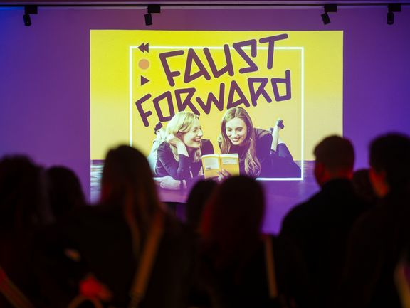 Hinter einer verschwommenen Menschengruppe im Vordergrund ist das gelbe Coverbild des Podcasts "Faust Forward" an die Wand projiziert. 