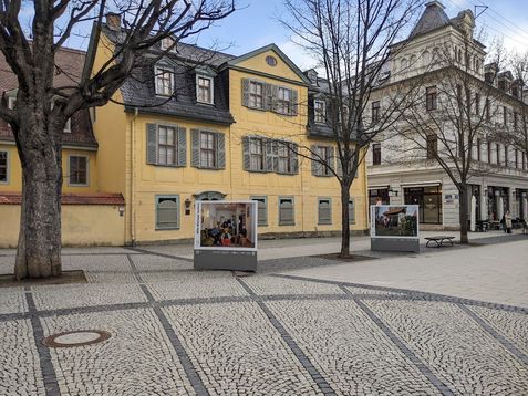 Dreiecksaufsteller mit einer Fotoserie, die das Wohnen in Weimar zeigt, steht vor einem gelben Haus
