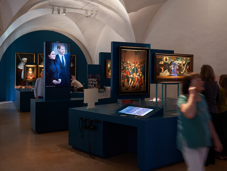 Ausstellungsraum mit Gewölbedecke, in dem verschiedene Gemäde hängen und sich Menschen bewegen.